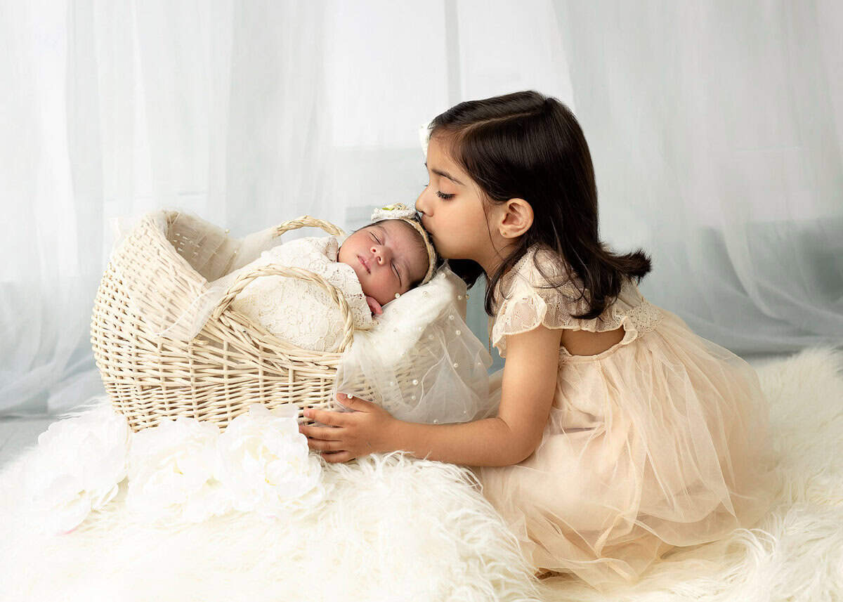 newborn photography of big sister kissing baby tucked in a bucket with white linen